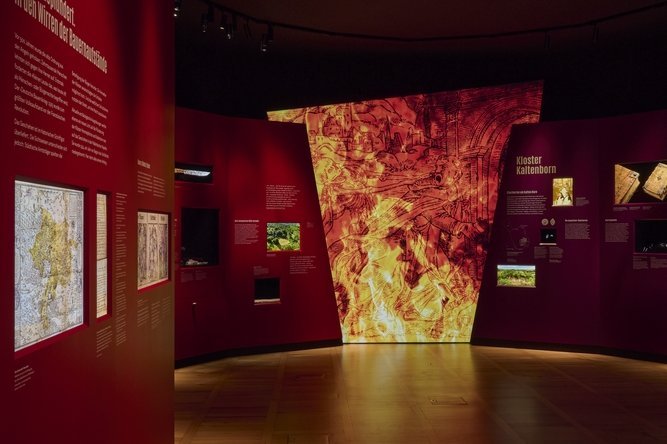 Blick in die Ausstellung: Zwischen den Themen Kloster Himmelpforte (links in Rot) und Stift Kaltenborn (rechts in Purpur) lodern Flammen. Die Ausstellungsgestaltung des halleschen Künstlers Klaus Pockrandt schuf ein Sinnbild für den Untergang großer Teile der mittelalterlichen Klosterlandschaft in den Aufständen des Jahres 1525. © Landesamt für Denkmalpflege und Archäologie Sachsen-Anhalt, Thomas Puttkammer.